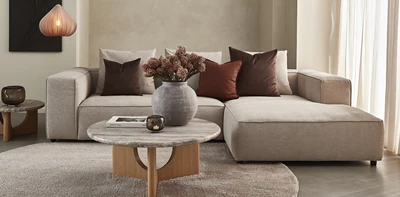 Elegant beige hjørnesofa med pyntepuder i varme nuancer. Et rundt marmorbord med træben og en vase med tørrede blomster fuldender indretningen