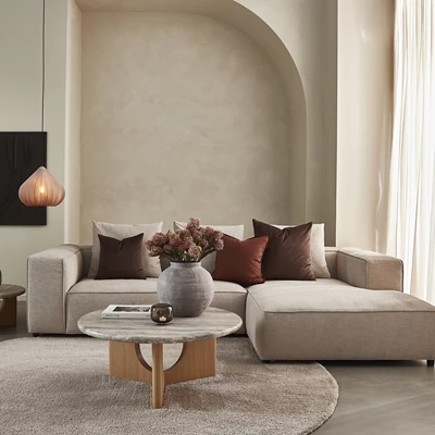 Elegant beige hjørnesofa med pyntepuder i varme nuancer. Et rundt marmorbord med træben og en vase med tørrede blomster fuldender indretningen