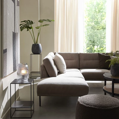 Udsnit af stue, hvor en sofa med puf i gråt betræk er i centrum. Rundt om er bl.a. to sideborde med hhv. en lampe og en plante i sort krukke